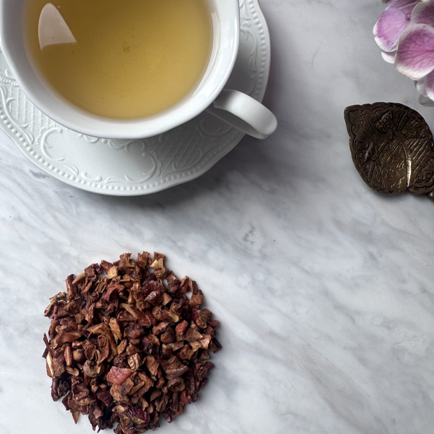 A delicate white teacup filled with pale golden Turkish Apple tea rests on a white saucer atop a marble surface. Beside it lies a small heap of the dried apple tea blend, while soft lavender hydrangeas frame the composition, creating an elegant and tranquil tea moment.