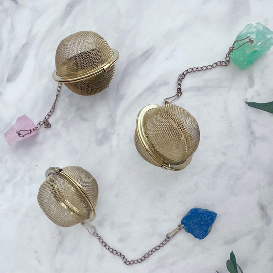 Three gold mesh tea infusers on a marble surface with decorative stones and leaves.